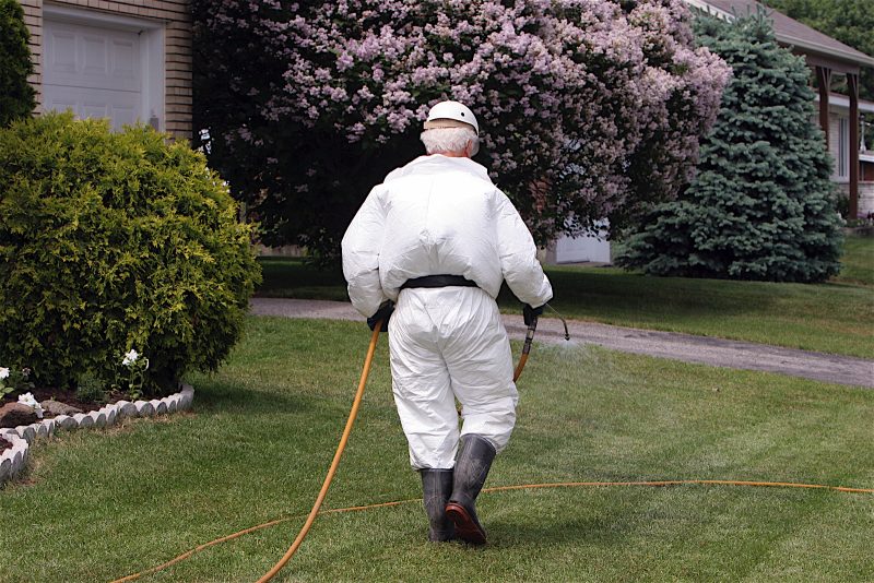 Pesticide technician walking on mowed lawn and dragging a long yellow hose behind him. He's fully covered by a white uniform, high black boots, black gloves and white hat.